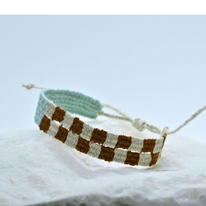 Op de afbeelding: Geweven armband met een geblokt patroon in bruin, wit en lichtblauw. De verstelbare armband is gemaakt van geweven stof en heeft een neutrale sluiting met een touwtje. De armband wordt gepresenteerd op een wit oppervlak.