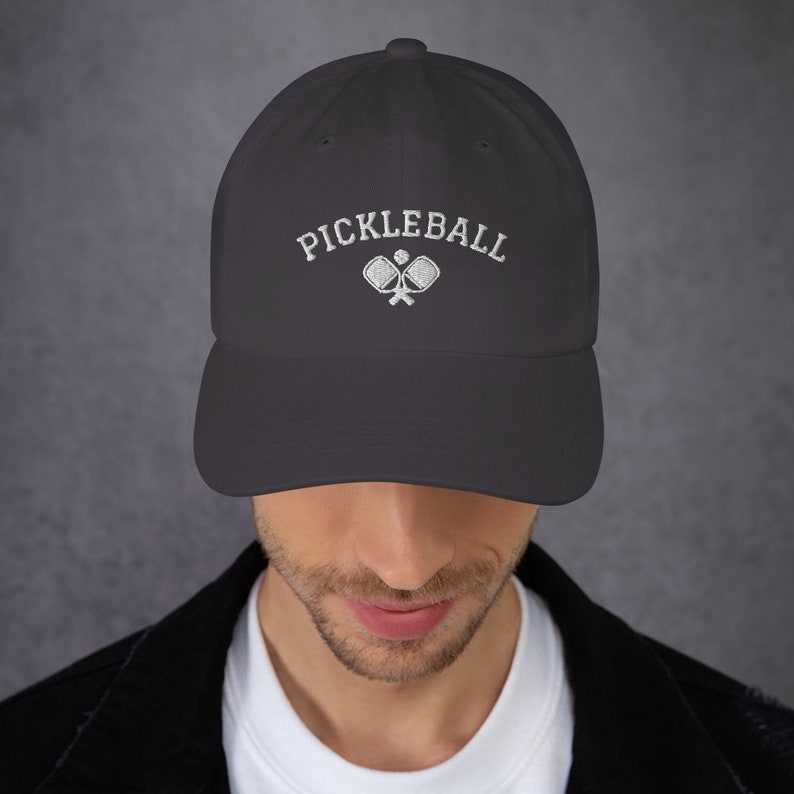 May include: A dark gray baseball cap with white embroidery that reads "Pickleball" and features a pair of crossed pickleball paddles.