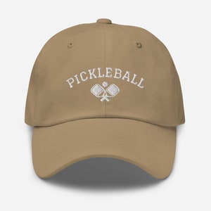 May include: A tan baseball cap with white embroidery that reads "Pickleball" with a graphic of two pickleball paddles and a ball.