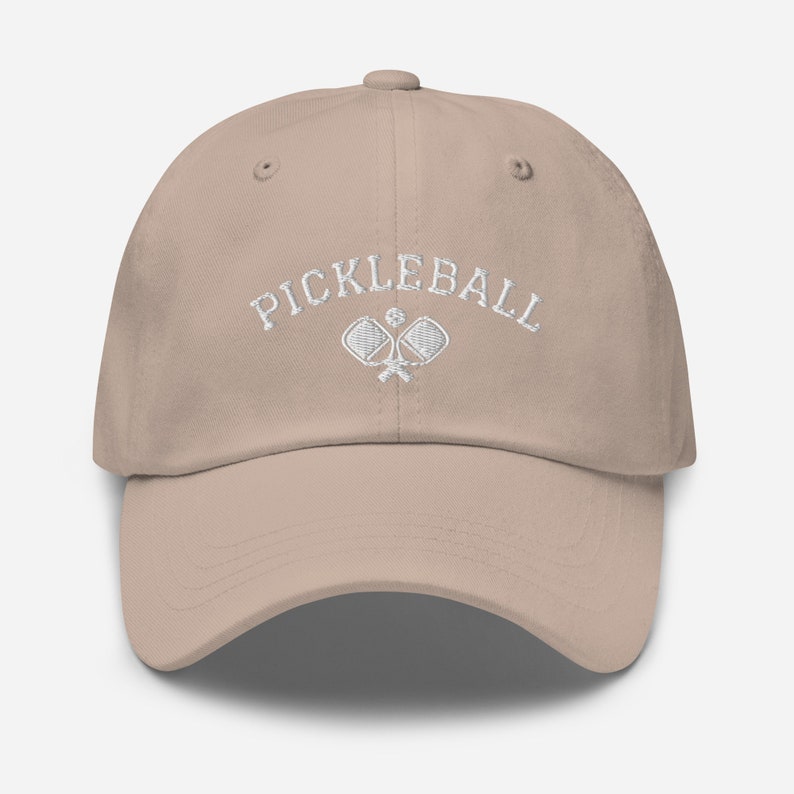 May include: A beige baseball cap with white embroidery that reads "Pickleball" and features two crossed pickleball paddles.