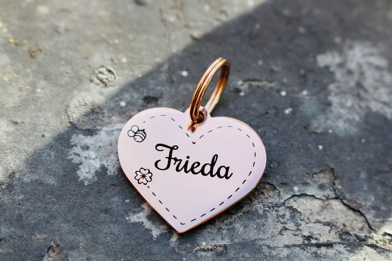 以下が含まれることがあります： 「Frieda」と刻まれた、ローズゴールドのハート型のペットタグ。タグには、小さな蜂と花のデザインが施されています。