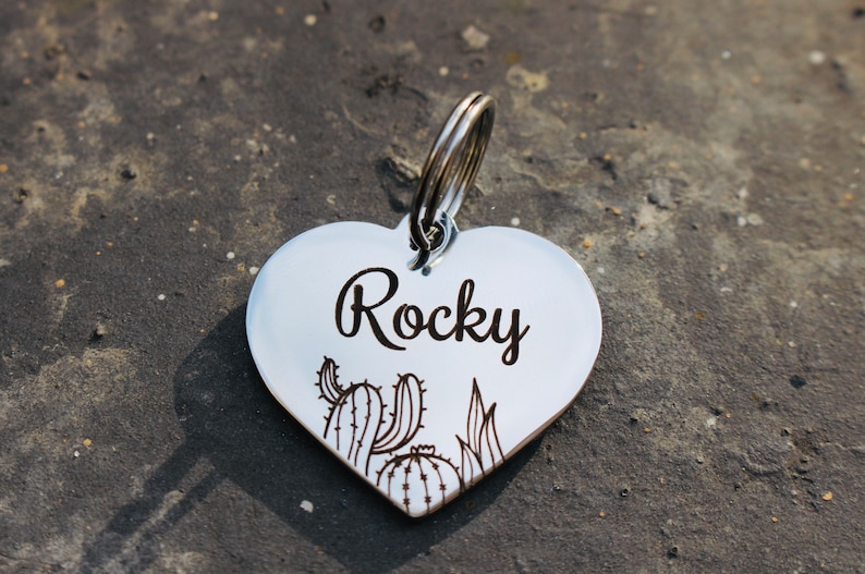 以下が含まれることがあります： 「ロッキー」と刻まれた銀色のハート型の犬用タグ。タグにはサボテンと多肉植物のデザインが施されています。