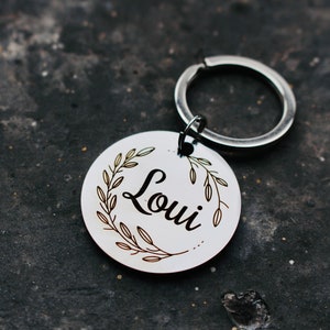May include: A silver key chain with a round tag that says "Loui" in a cursive font. The tag is surrounded by a wreath of leaves.