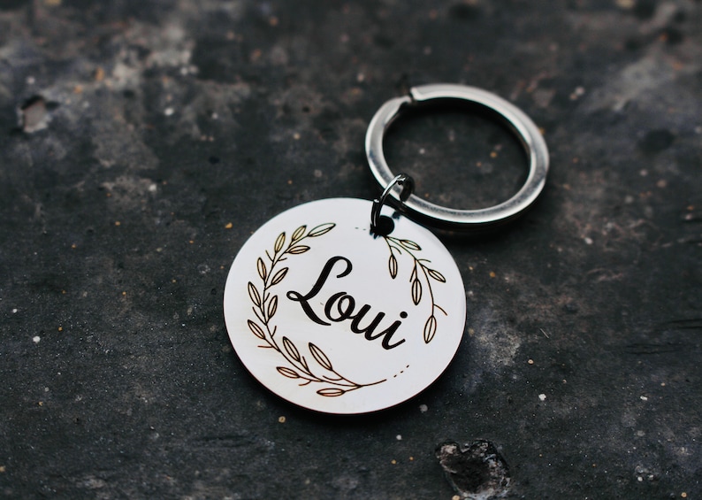 May include: A silver key chain with a round tag that says "Loui" in a cursive font. The tag is surrounded by a wreath of leaves.