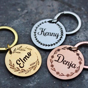 May include: Three personalized keychains with different names, "Elmo", "Kenny", and "Denja", engraved on round metal tags. Each tag features a decorative floral wreath design. The keychains are gold, silver, and rose gold.