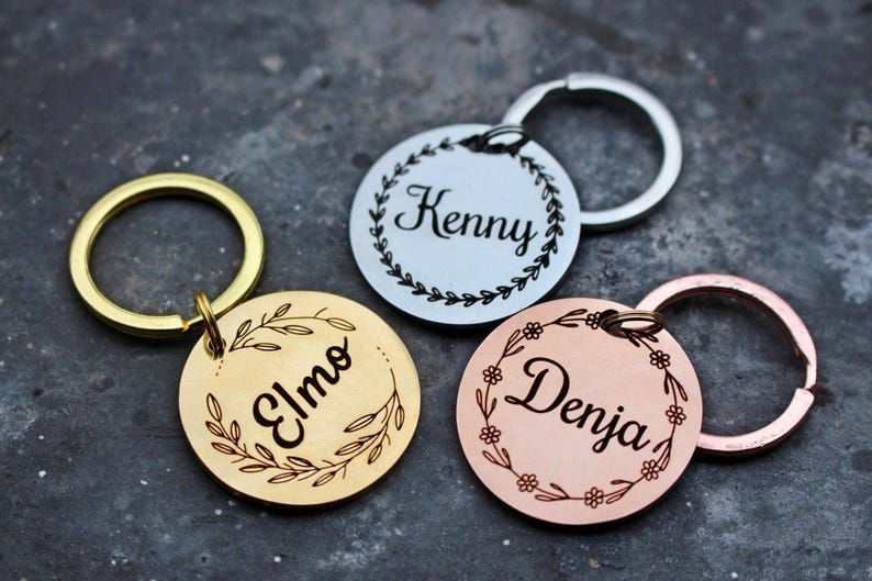 May include: Three personalized keychains with different names, "Elmo", "Kenny", and "Denja", engraved on round metal tags. Each tag features a decorative floral wreath design. The keychains are gold, silver, and rose gold.