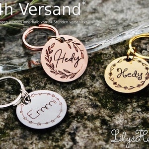 May include: Three personalized dog tag keychains with different designs. The keychains are made of metal and have a round shape. The first keychain has a copper finish with a wreath design and the name "Hedy" engraved on it. The second keychain has a gold finish with a floral design and the name "Hedy" engraved on it. The third keychain has a silver finish with a heart design and the name "Emmi" engraved on it. The keychains are displayed on a textured surface.