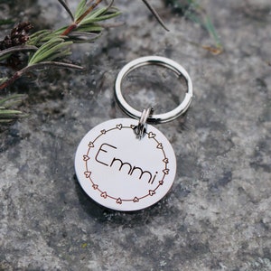 May include: Silver dog tag with a heart border and the name "Emmi" engraved on it. The tag is attached to a silver key ring.