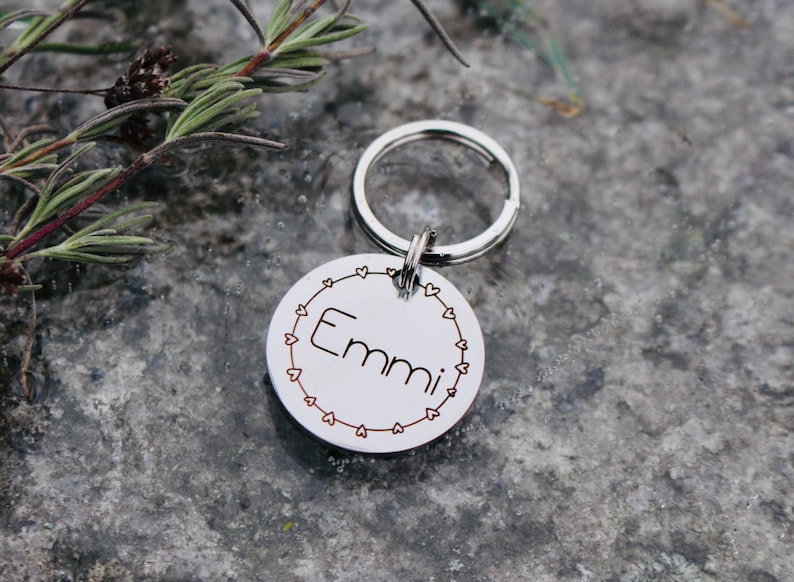 May include: Silver dog tag with a heart border and the name "Emmi" engraved on it. The tag is attached to a silver key ring.