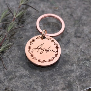May include: Rose gold keychain with a round tag engraved with the name "Ayoka" surrounded by a floral wreath.