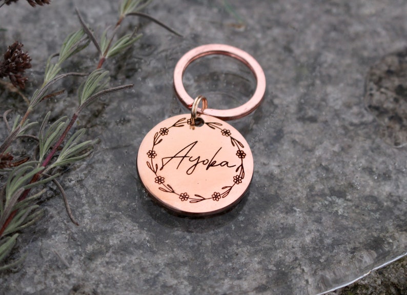 May include: Rose gold keychain with a round tag engraved with the name "Ayoka" surrounded by a floral wreath.