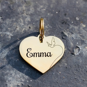 以下が含まれることがあります： 小さな鳥のデザインと「Emma」という名前が刻まれた、金色のハート型のペットタグです。