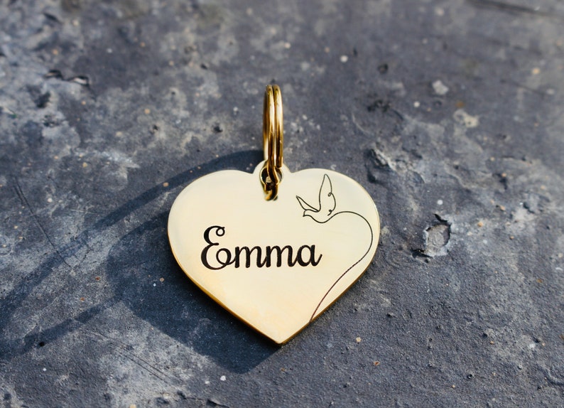 以下が含まれることがあります： 小さな鳥のデザインと「Emma」という名前が刻まれた、金色のハート型のペットタグです。