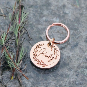 May include: Rose gold key chain with a round tag engraved with the name "Hedy" surrounded by a wreath of leaves.