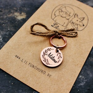 May include: A copper dog tag with a wreath design and the name "Milou" engraved on it. The tag is attached to a silver key ring and a piece of twine. The tag is on a brown paper card with the website "www.lilyundhund.de" printed on it.
