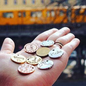 May include: A hand holding eight round metal tags with engraved names, including "Honey", "Iskra", "Raoul", "Runa", "Buffy", "Kalle", and two others. The tags are in various colors, including gold, silver, and rose gold.