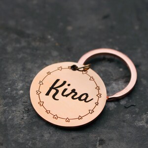 May include: A rose gold keychain with a round tag that says "Kira" in a cursive font. The tag is surrounded by a circle of small hearts.