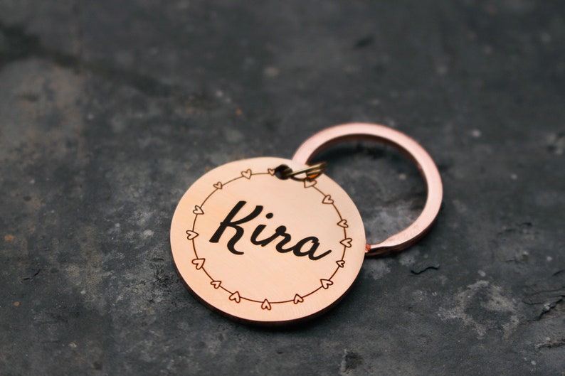 May include: A rose gold keychain with a round tag that says "Kira" in a cursive font. The tag is surrounded by a circle of small hearts.