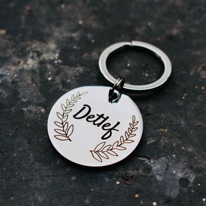 May include: Silver keychain with a round tag featuring a leafy wreath design and the name "Detlef" engraved in black.