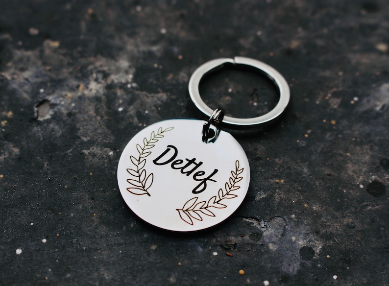 May include: Silver keychain with a round tag featuring a leafy wreath design and the name "Detlef" engraved in black.