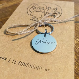 May include: A silver dog tag with the name "Wilson" engraved on it. The tag is attached to a silver key ring and a brown twine string. The tag is sitting on a brown paper bag with the website "www.lilyundhund.de" printed on it.