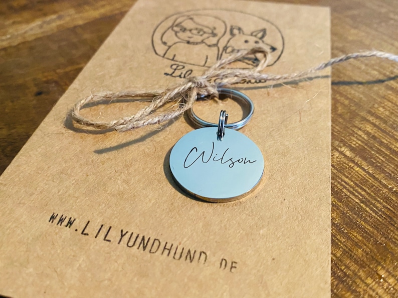May include: A silver dog tag with the name "Wilson" engraved on it. The tag is attached to a silver key ring and a brown twine string. The tag is sitting on a brown paper bag with the website "www.lilyundhund.de" printed on it.