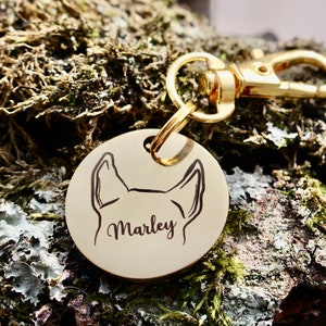 Puede incluir: Chapa de perro dorada con un contorno negro de la cabeza de un perro y el nombre "Marley" grabado en ella.