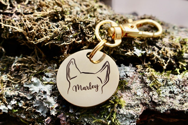 Puede incluir: Chapa de perro dorada con un contorno negro de la cabeza de un perro y el nombre "Marley" grabado en ella.