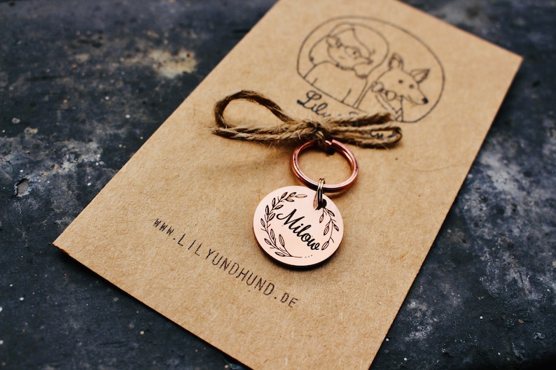 May include: A copper dog tag with a wreath design and the name "Milou" engraved on it. The tag is attached to a silver key ring and a brown twine string. The tag is on a brown paper card with the website "www.lilyundhund.de" printed on it.