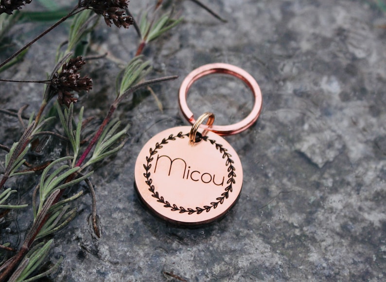 May include: A round copper-colored dog tag with a key ring. The tag has a wreath design around the name "Micou" engraved in the center.