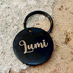 May include: Black round dog tag with a gold engraved name "Lumi" and a small heart. The tag is attached to a black metal ring.