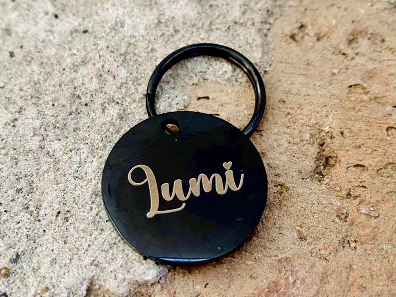 May include: Black round dog tag with a gold engraved name "Lumi" and a small heart. The tag is attached to a black metal ring.
