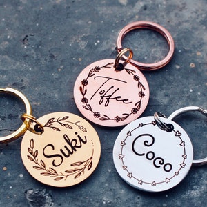 May include: Three personalized dog tags with engraved names "Suki", "Toffee", and "Coco". The tags are round and have different colored metal finishes: gold, rose gold, and silver. Each tag has a decorative border with floral or heart designs.