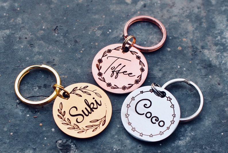 May include: Three personalized dog tags with engraved names "Suki", "Toffee", and "Coco". The tags are round and have different colored metal finishes: gold, rose gold, and silver. Each tag has a decorative border with floral or heart designs.