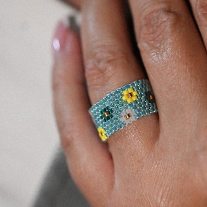 Può includere: Un anello di perline con sfondo turchese e motivo floreale. I fiori sono gialli, bianchi e verdi. L'anello è indossato su un dito.
