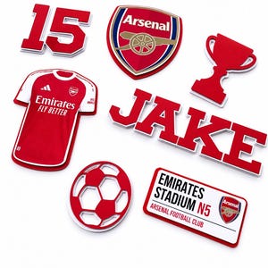 Puede incluir: Una colección de artículos rojos y blancos con el tema del club de fútbol Arsenal. Incluye una camiseta, un balón de fútbol, un trofeo y el nombre "JAKE". También presenta el número 15, el logotipo del club y un letrero.
