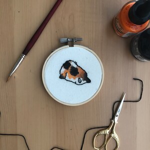 May include: A white embroidery hoop with a calico cat design. The cat is curled up and sleeping. The hoop is on a wooden surface.