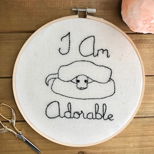 May include: Embroidered hoop art with a black and white illustration of a snake curled up with the text "I am adorable" in black thread.