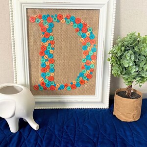 May include: A white framed picture with a burlap background and the letter D made out of colorful buttons. The buttons are in shades of orange, blue, and green. There is a white ceramic elephant planter in the foreground and a small potted green plant to the right of the picture.