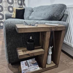 May include: A stained wooden side table with two shelves. The table holds a black jar, a small white statue, and books. A pair of glasses and a remote control rest on the top surface. The table is next to a grey sofa.