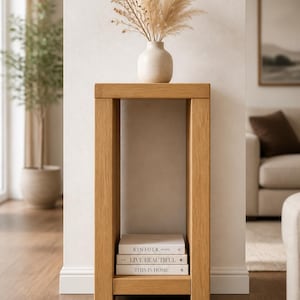 May include: A rectangular light oak wood console table with a flat top and a lower shelf. The table features a clean, block-style frame. On the top sits a beige ceramic vase with dried pampas grass. Three books are stacked on the bottom shelf.