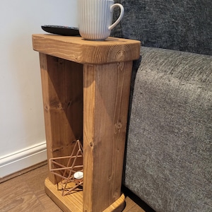 May include: A tall, narrow wooden side table with a medium brown finish and visible wood grain. The table features a rectangular top and base connected by two vertical supports. A white ribbed mug and a black remote control sit on the top surface.