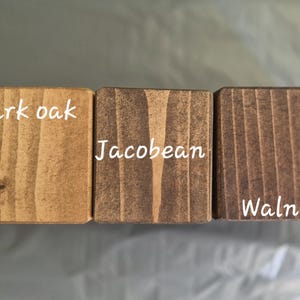 May include: Three wooden blocks with different stain colors: Dark Oak, Jacobean, and Walnut. Each block has the stain name in white text. The wood grain is visible, showcasing the natural texture and color variations of the wood.