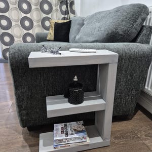 May include: A gray, C-shaped side table with two shelves. A black decorative jar and two books are on the shelves. A pair of glasses and a remote control are on the top shelf. The table is next to a gray sofa.