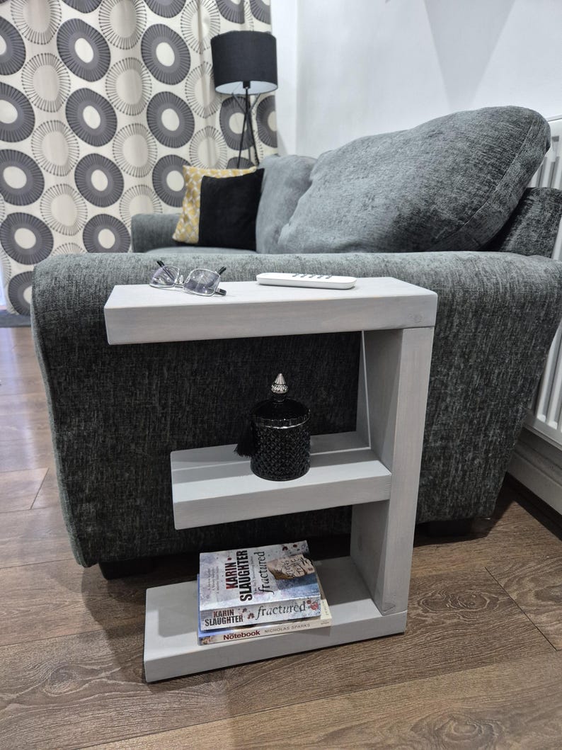 May include: A gray, C-shaped side table with two shelves. A black decorative jar and two books are on the shelves. A pair of glasses and a remote control are on the top shelf. The table is next to a gray sofa.