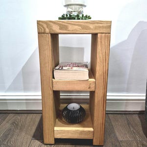 May include: A rectangular wooden side table with a light brown finish, featuring a flat top surface and one middle shelf. The table is constructed from thick, solid wood planks. A book rests on the middle shelf, and a small black bowl sits below.