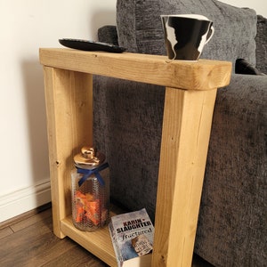 May include: A natural wood side table with a shelf. A black remote, a black and white mug, a glass jar with a gold lid and blue ribbon, and a book titled "Fractured" are on the table. The table is beside a grey sofa.
