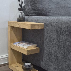 May include: A light-colored wooden side table with two shelves. A book, a candle, and a glass diffuser bottle with black reeds are on the shelves. The table is next to a gray sofa.