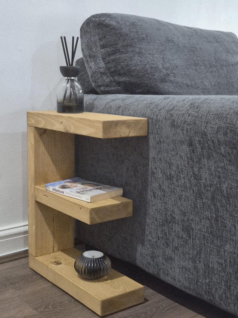 May include: A light-colored wooden side table with two shelves. A book, a candle, and a glass diffuser bottle with black reeds are on the shelves. The table is next to a gray sofa.