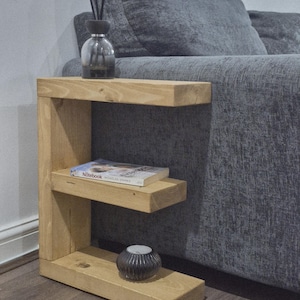 May include: A light-colored wooden side table with two shelves. A book, a candle holder, and a diffuser bottle with reeds are on the shelves. The table is next to a gray sofa.